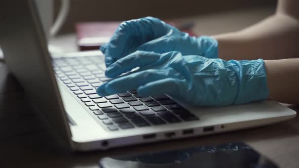 A Woman in Disposable Gloves and a Mask Works at Home with a Laptop