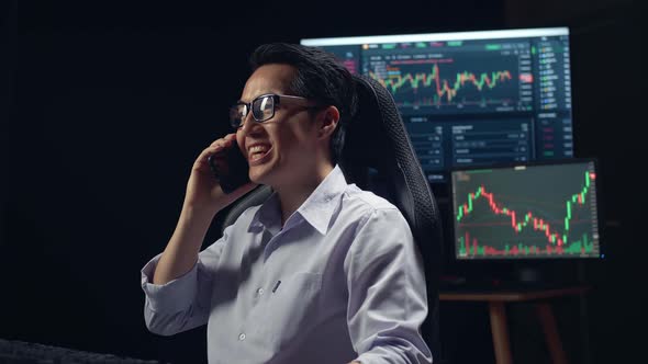 Asian Young Male Stock Market Broker Talking On Mobile Phone With Analyzing Graphs