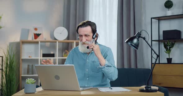 Mature Man in Headset which Sitting in front of Computer at Home During Video Meeting