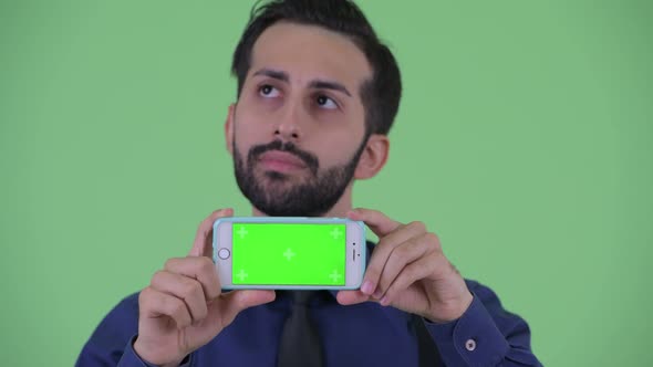 Face of Happy Young Bearded Persian Businessman Thinking While Showing Phone
