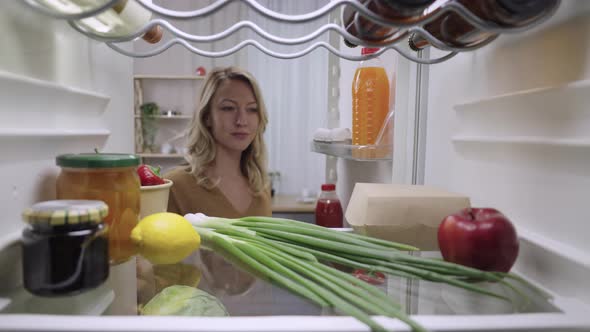 Young Woman Opens the Refrigerator Door and Takes Out a Burger Box and Juice