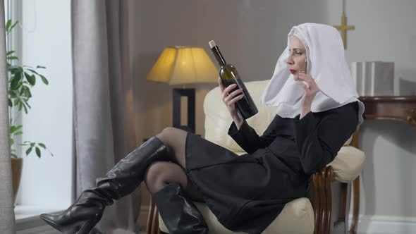 Side View of Concentrated Confident Woman in Nun Costume Sitting on Armchair Examining Wine Bottle