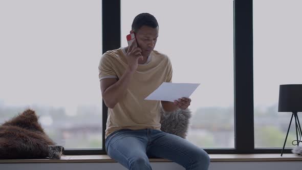 Portrait of African American Male Analyst Examining Graph Talking on the Phone Sitting on Windowsill
