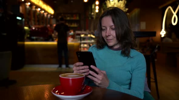 Cheerful Woman Is Resting Alone in Cafeteria and Reading Message in Her Smartphone, Smiling and