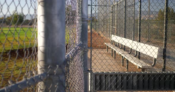 Slow pan across a chain link fence and an empty baseball field bench in the dugout of a local park a