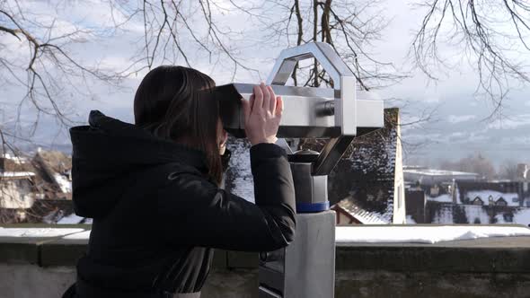 Young woman uses metal fixed coin-operated binoculars to survey a snow-covered wintry small town in