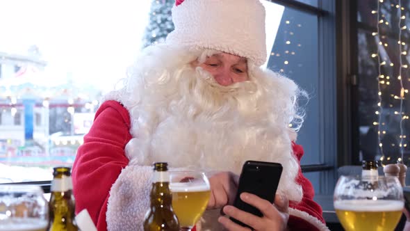 Santa Claus with a Smartphone in His Hands Drinks Beer and Writes Text Messages on Social Networks