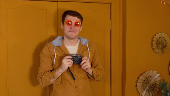 Young Man in Orange Glasses with Old Photo Camera Stands Against Background of Brown Doors in Room