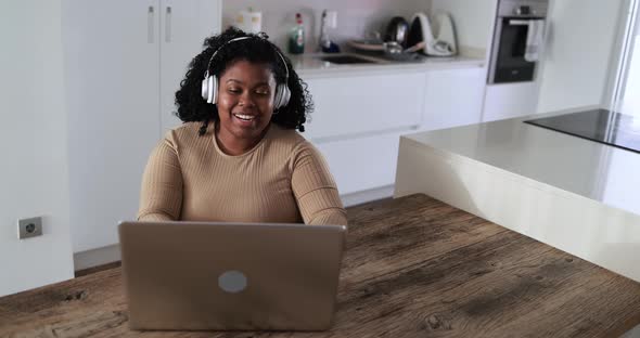 Happy african girl sitting inside kitchen at home using laptop computer while wearing headphones