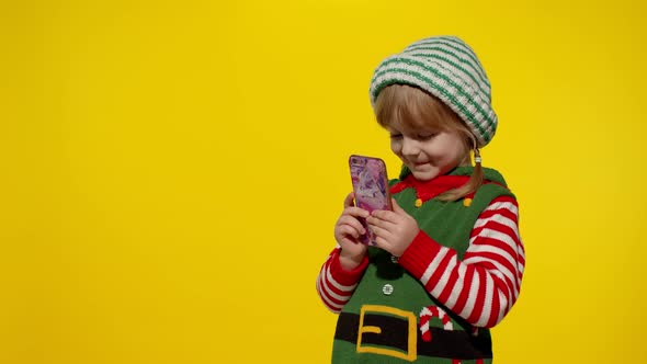 Kid Girl with Mobile Phone in Christmas Elf Santa Helper Costume Points Fingers Aside on Blank Wall