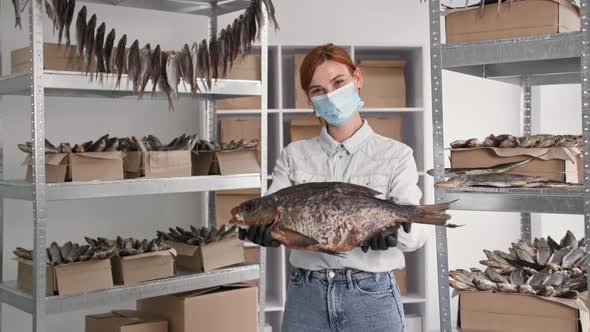 Portrait of Young Woman in Medical Mask with Large Sea Fish in Her Hands Background of Shelves with