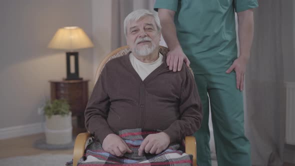 Portrait of Elderly Caucasian Man Talking at Camera As Unrecognizable Male Nurse Holding Hand on His