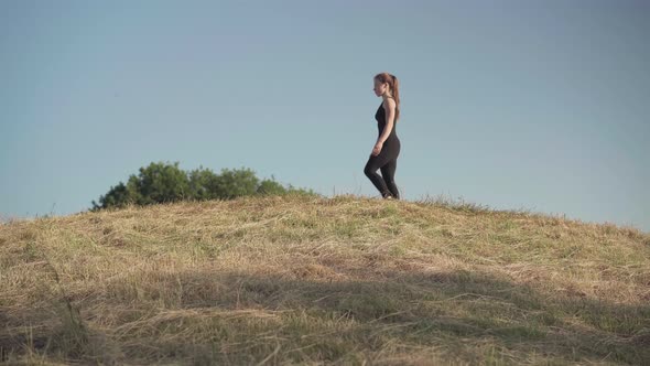 Wide Shot of Caucasian Female Gymnast Walking Along Hill and Jumping Up Raising Leg Up. Side View