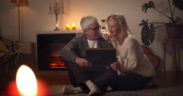 Senior Couple Looking at Framed Family Photo Sitting on Floor Near Fireplace