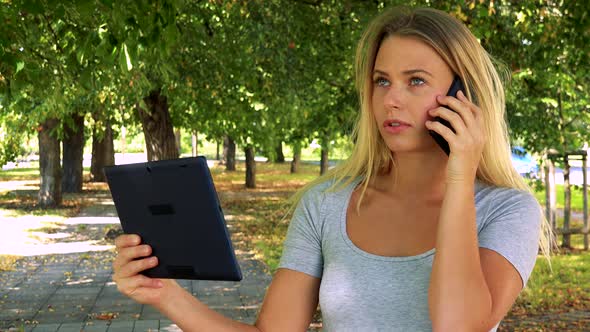 Young Pretty Blond Woman Works on Tablet and Phone with Smartphone - Park with Trees in Background