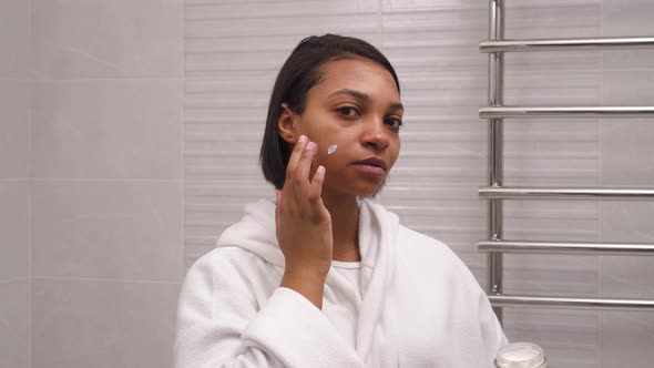 A Woman with Dark Skin Uses a Facial Moisturizer in the Morning in the Bathroom