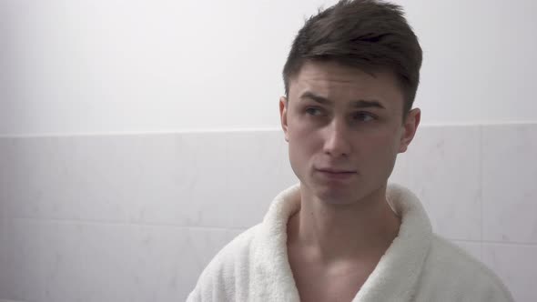 Attractive Young Man in a White Bathrobe Standing in Front of a Bathroom Mirror and Fixing His Hair