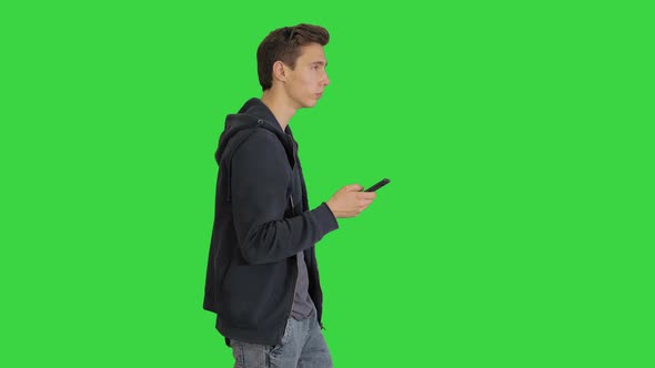 Young Man in Casual Clothes Walking and Texting on Cell Phone While Walking on a Green Screen