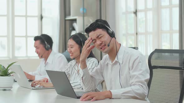 A Man Of Three Asian Call Centre Agents Wearing Headset And Tired Due To Working At The Office