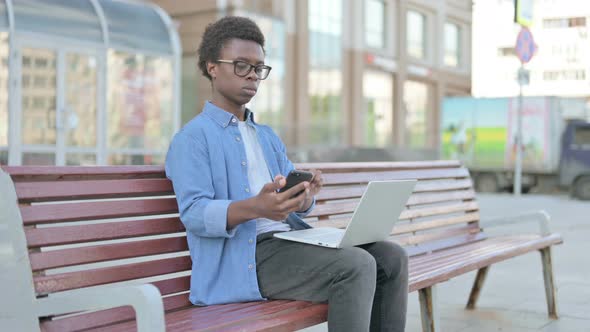 Young African Man Using Smartphone and Laptop While Sitting Outdoor on Bench