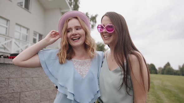 One Young Girl in a Hat and Another in Funny Glasses Fool Around in Front of the Camera and Grimace