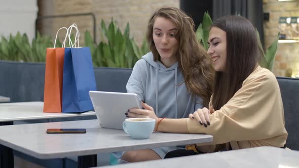 Two Young Positive Millennial Women Talking and Laughing Sitting in Cafe with Tablet and Shopping