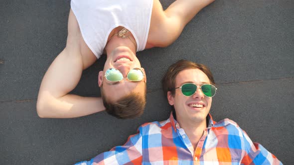 Young Happy Male Couple in Sunglasses Lying on Rooftop of High-rise Building and Candid Laughing