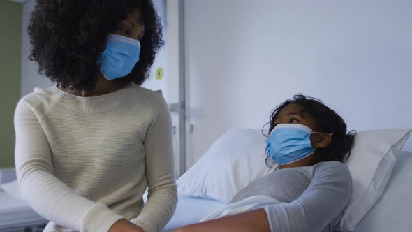 African american mother wearing face mask holding hands and comforting her daughter at hospital