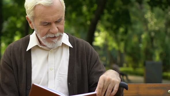 Old Wrinkled Man With Cane Sitting on Bench and Reading Book at Green Park