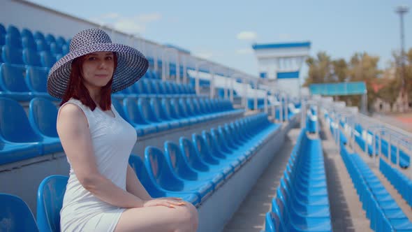 Young woman in white dress and elegant hat looks at camera, sitting on stadium bleachers alone.