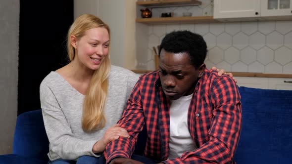 Caucasian Woman Calms African American Upset Man Sitting on Couch at Home