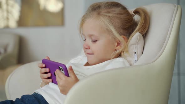 Little Blonde Girl is Watching a Video on Her Phone While Sitting in a Chair