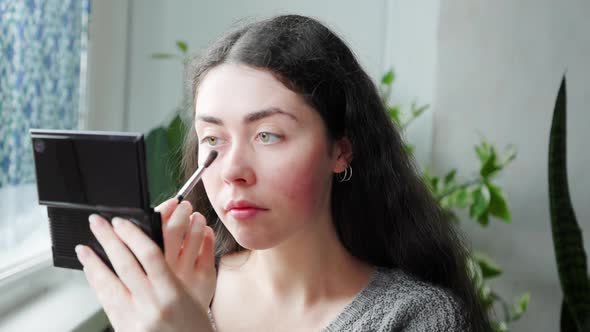 Portrait of a Young Beautiful Woman Applying Makeup at Home Using a Brush and a Compact Mirror