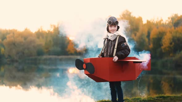 Happy Little Aviator Girl Standing at Sunset Lake in Cardboard Plane Costume with Color Smoke