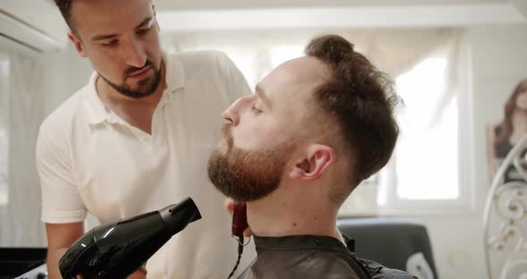 The Hairdresser Works with a Hair Dryer and Combs a Beard