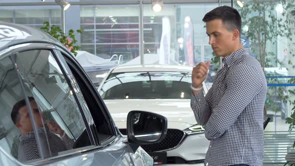 Young Man Rubbing His Chin Thoughtfully, Choosing New Car To Buy at Dealership