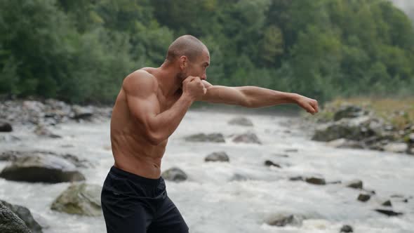 a Man with a Naked Torso is Boxing By a Mountain River