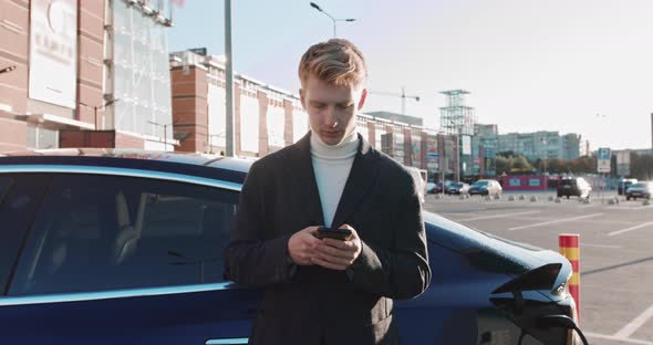 Businessman With A Smartphone In His Hands Leans On A Blue Luxury Electric Car