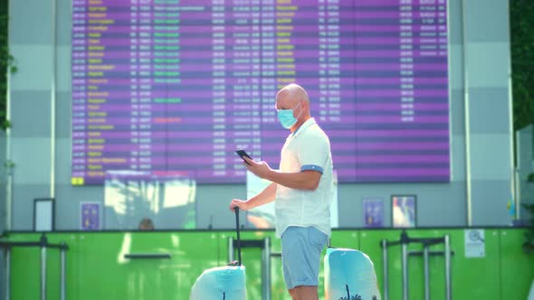 Male Air Passenger in Medical Mask, with Luggage, Standing Against Departure Board at Airport
