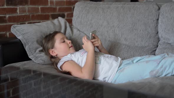 Little girl lying on couch and looking at smartphone at home indoors. Child plays and watching video