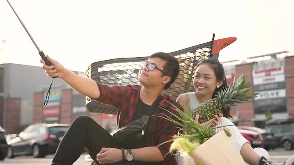 Happy Vietnamese Couple Sitting on the Ground Near Big Shopping Centre and Making Selfie