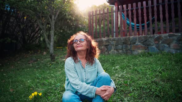 A Mature Adult Woman in Glasses Sits in a Country House's Garden