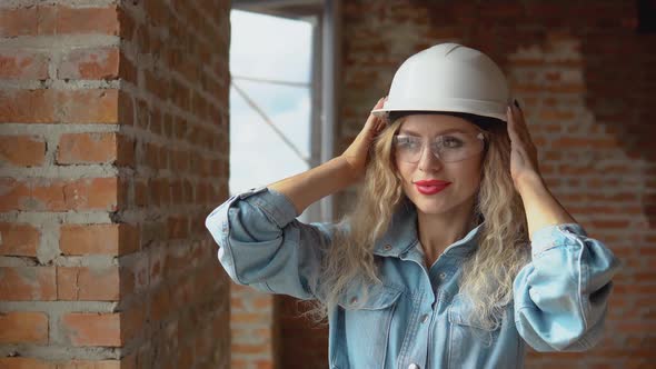 A Young Woman in a Work Helmet and Denim Clothes and Goggles Stands at the Construction Site