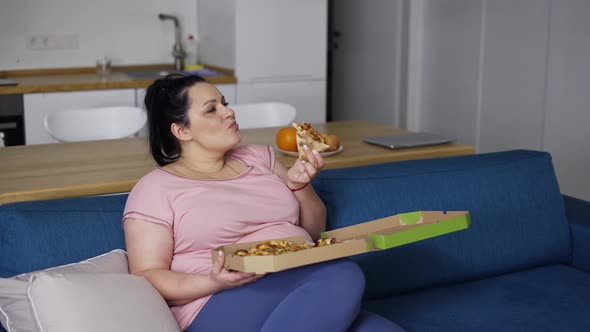 Plus Size Woman Eats Slice of Pizza with Great Pleasure Sitting on a Couch