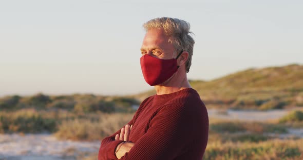 Portrait of happy caucasian man in face mask standing with arms crossed on sunny day at the beach