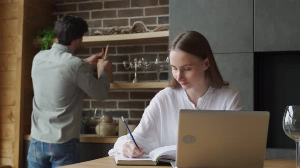 Young Woman Using a Laptop in the Kitchen and a Man Prepares Food