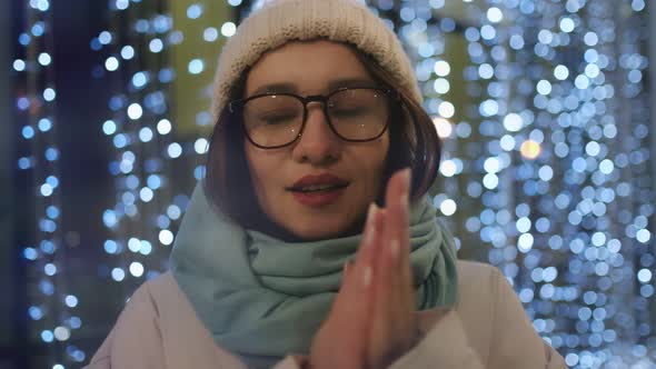 Beautiful brunette in glasses and a winter hat against the background of a garland. Waiting concept