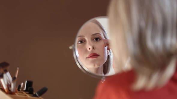 Beautiful blond hair woman in red shirt applying makeup in opposite of mirror