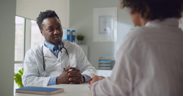 Afroamerican Male Doctor Talking to Patient Sitting at Desk in Office