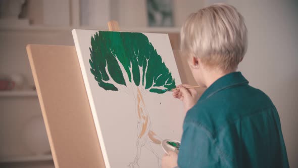 A Young Blonde Woman Draws a Painting with a Green Color on the Canvas